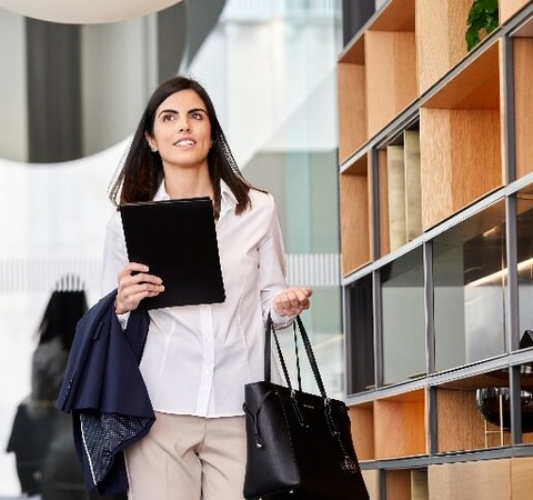A businesswoman walks through a modern office space carrying a tablet and a handbag.