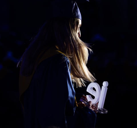 A graduate holds a sign while looking away in a dimly lit ceremony.