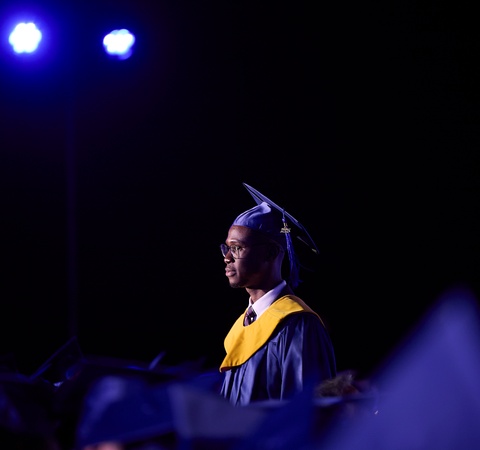 A graduate wearing a blue gown and yellow stole stands proudly during a graduation ceremony.