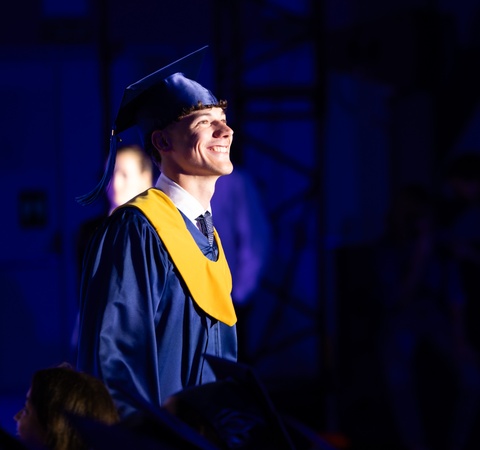 A smiling graduate stands proudly in a blue cap and gown.