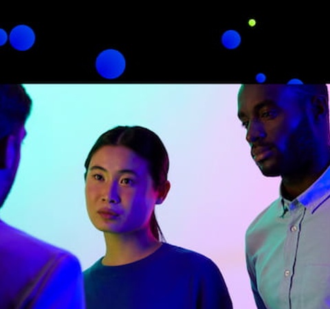 Three people, one woman and two men, engaged in a conversation in a room with colorful lighting.