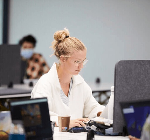 A woman with glasses working intently at a desk in a busy office environment.