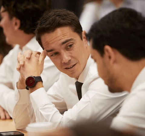 A man in a formal shirt and tie engaging in a serious discussion with another man at a crowded meeting table.