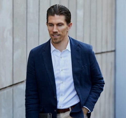 A man in a blue blazer walks casually along a textured wall.