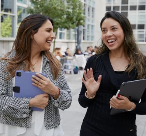 Two women are smiling and talking while holding tablets in an outdoor setting.