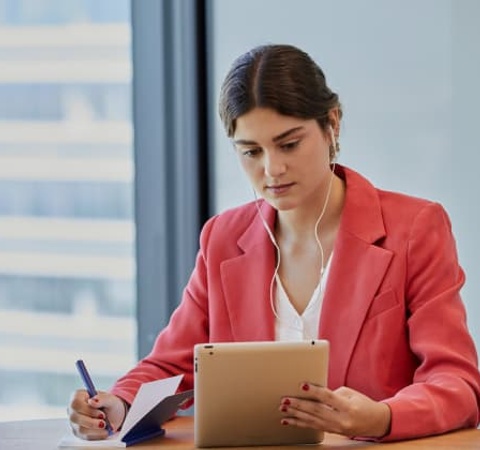 A woman in a red blazer is sitting at a table, working intently on a tablet while listening to music.