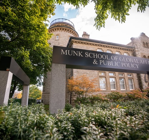 View of the Munk School of Global Affairs & Public Policy building behind a sign and lush greenery.