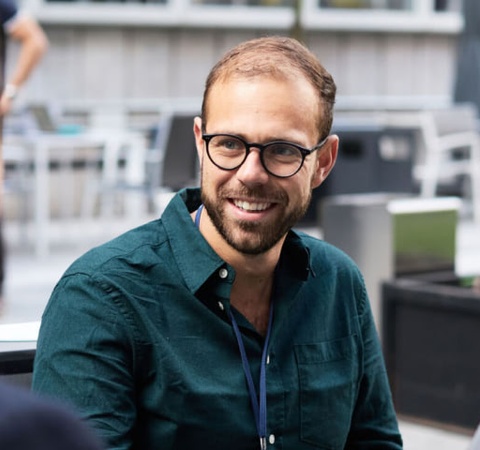 A man in glasses smiling during a conversation with another person at an outdoor table.