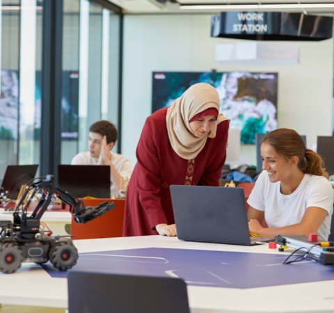 A woman in a hijab assists another woman at a laptop in a modern tech workspace filled with students and robotic equipment.