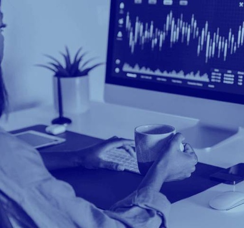 A woman analyzes financial data on a computer screen while holding a cup of coffee in an office setting.