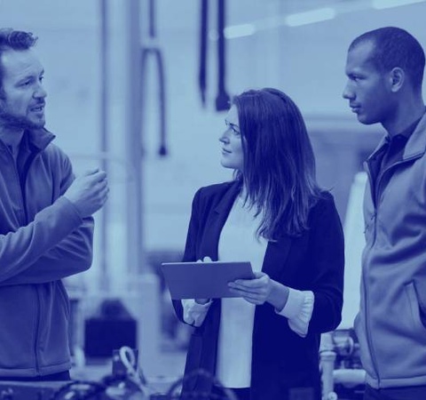 Three professionals discussing in an industrial setting, with one holding a tablet.