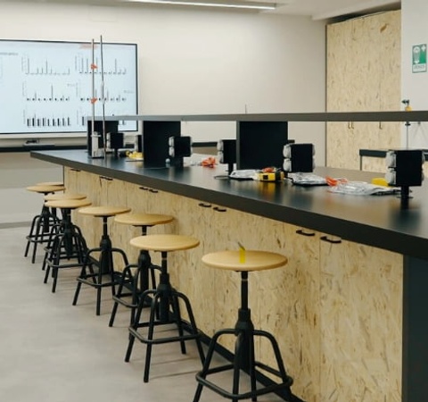 A modern laboratory with a long black countertop, stools, and a large screen displaying data.