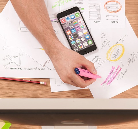 An overhead view of a person's hands working on a creative project with papers, pens, a smartphone, and stationery items on a wooden desk.