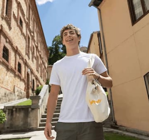 A young man in a white T-shirt smiles while walking outdoors near a building.