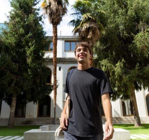 A young man smiling in a courtyard surrounded by palm trees and historic buildings.