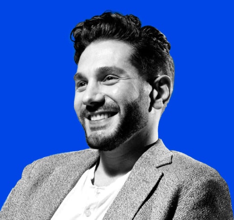 A portrait of a smiling man with black hair and a beard, wearing a grey blazer over a white shirt, set against a bright blue background.
