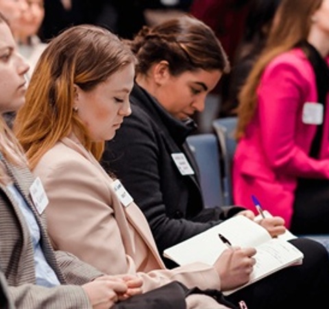 A group of people seated attentively during a conference or seminar, with some taking notes.