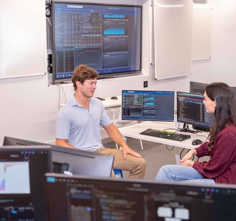 Two individuals are engaged in a discussion in a modern classroom equipped with multiple computer screens displaying data.