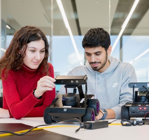 Two students are working together on a robotic device in a modern workspace.