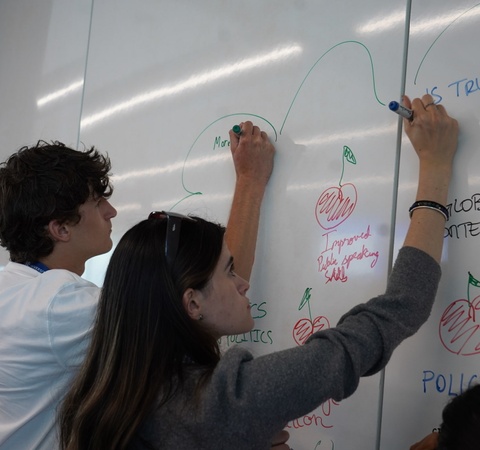Two individuals are writing on a whiteboard filled with colorful notes and drawings.