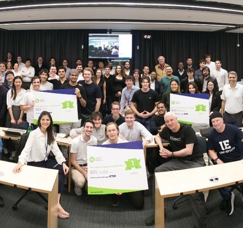 A group of people posing for a photo in a classroom, some holding large banners with logos.