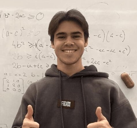 A young man smiles and gives a thumbs up in front of a whiteboard filled with mathematical equations.