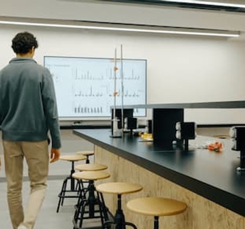 A man walking towards a whiteboard in a modern laboratory setting with stools and workbenches.