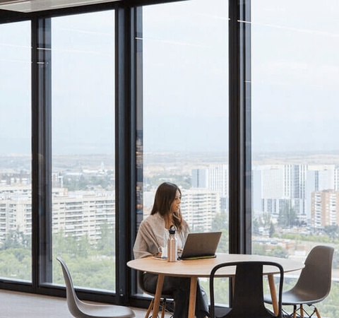 A woman works on a laptop at a table in a modern office with large windows overlooking a cityscape.