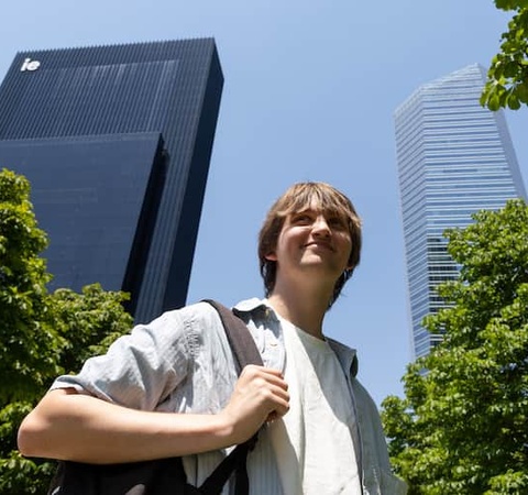 A young man stands in an urban area with modern skyscrapers and green trees around him.
