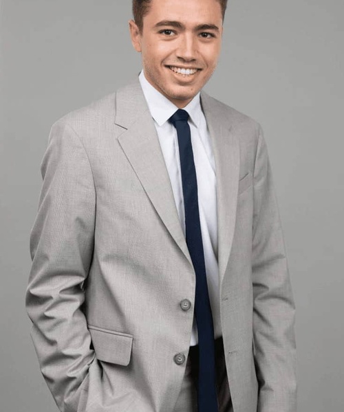A smiling man in a gray suit with a white shirt and blue tie standing against a gray background.