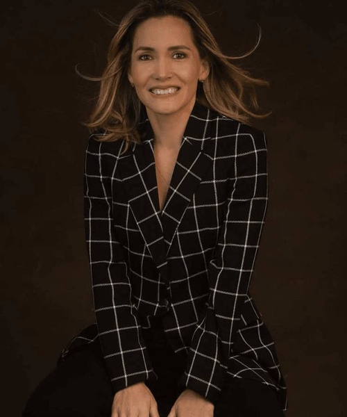A woman in a plaid blazer smiles while sitting against a dark background.