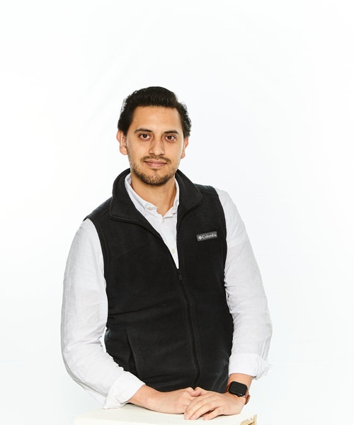 A confident young man in a white shirt and black vest poses against a white background.