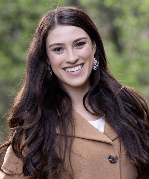 A smiling woman with long brown hair wearing a beige coat and diamond earrings outdoors.