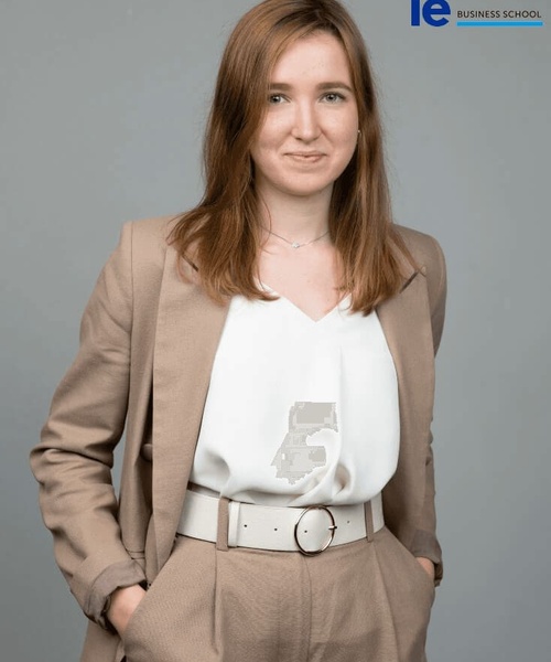 A young woman in a beige suit and white blouse stands against a grey background, labeled 'IE Business School'.