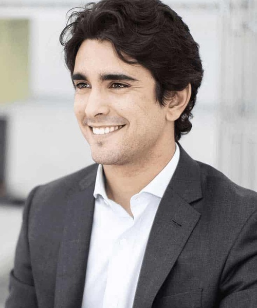 A smiling young man with dark curly hair wearing a gray suit and white shirt in an office environment.