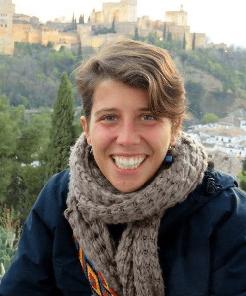 A smiling woman with short hair wearing a scarf, with the Alhambra in the background.
