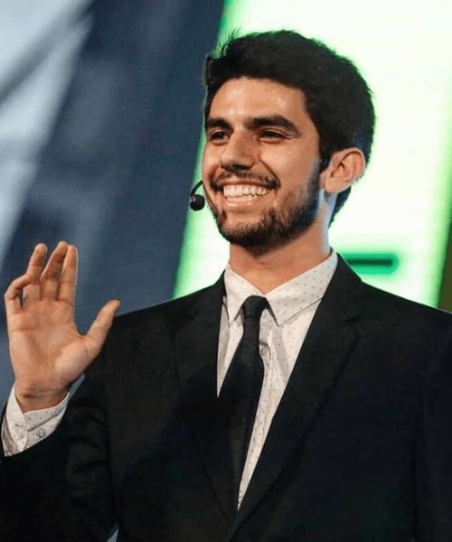 A young man in a suit is smiling and waving, with a microphone headset on, standing on a stage.