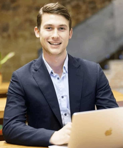 A young businessman smiling at the camera while working on his laptop in an office setting.