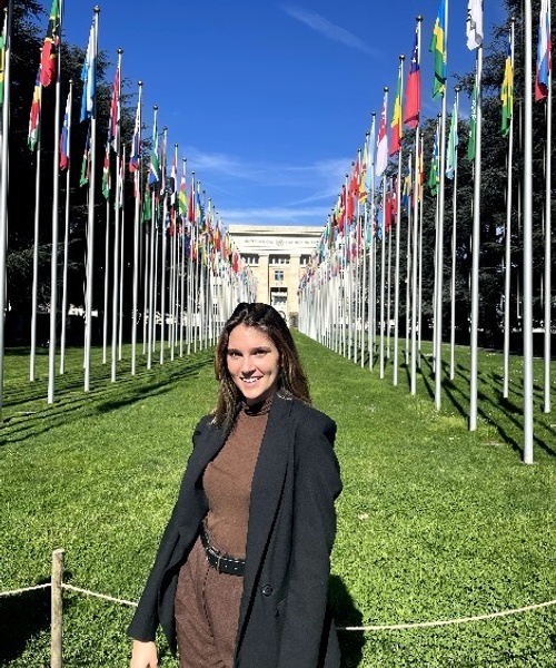 A woman smiling in front of a row of international flags outside a building.