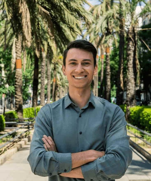 A smiling young man with arms crossed standing in a park with palm trees in the background.