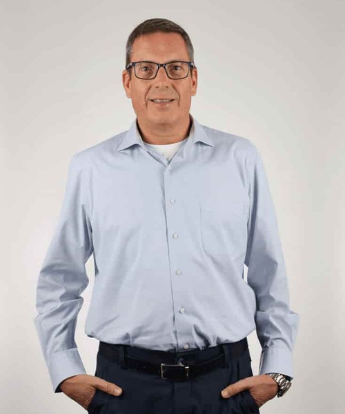 A man in a light blue shirt and black pants standing with his hands on his hips against a gray background.