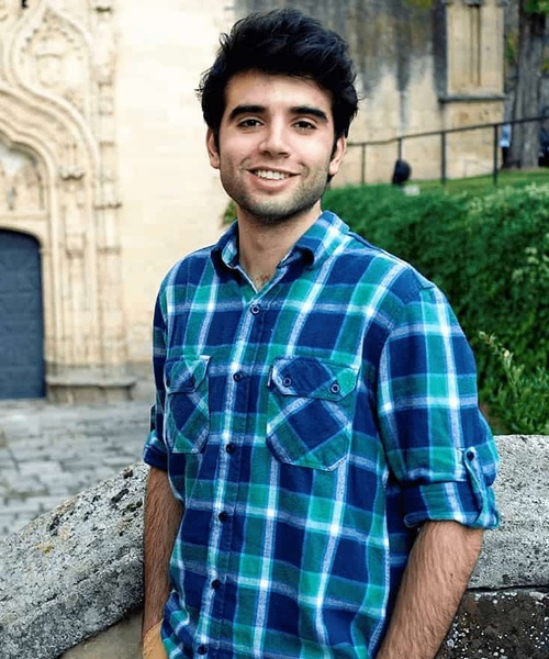 A young man in a blue plaid shirt smiling at the camera outdoors with an old stone building in the background.