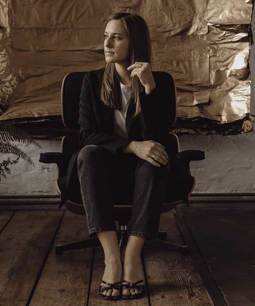 A woman in a thoughtful pose sits on a chair, dressed in a dark blazer and jeans, against a rustic backdrop.