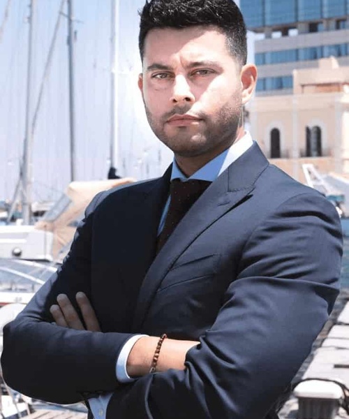 A confident man in a business suit stands with his arms crossed at a marina.