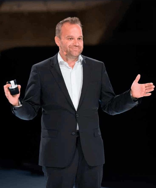 A man in a black suit and white shirt, standing with arms spread, holding a small object in one hand, and smiling while standing on a stage.