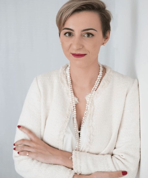 A professional woman in a white jacket and pearl necklace is posing with crossed arms against a light background.