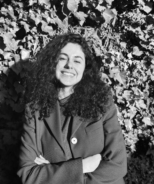 A black and white photo of a smiling woman with curly hair, standing in front of a leafy background.
