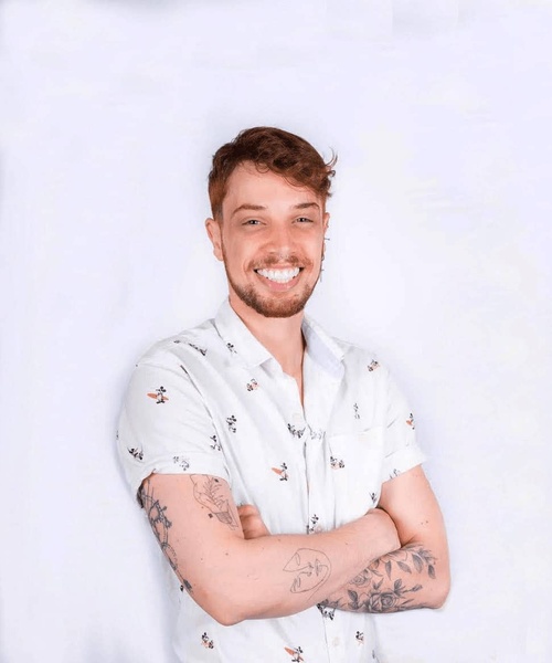 A young man with a beard, tattoos, and a floral shirt smiling confidently at the camera.