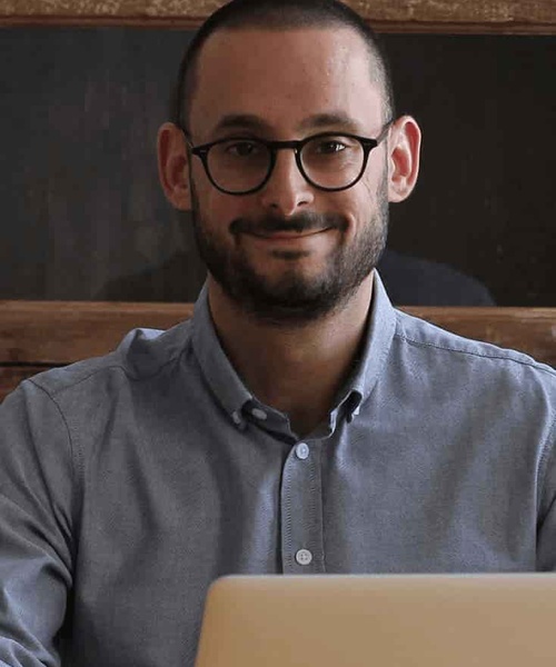 A man with glasses sitting in front of a laptop, smiling at the camera.