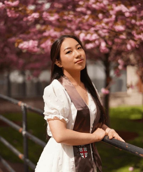 A young woman in a white dress stands by a railing, with blooming pink cherry blossoms in the background.